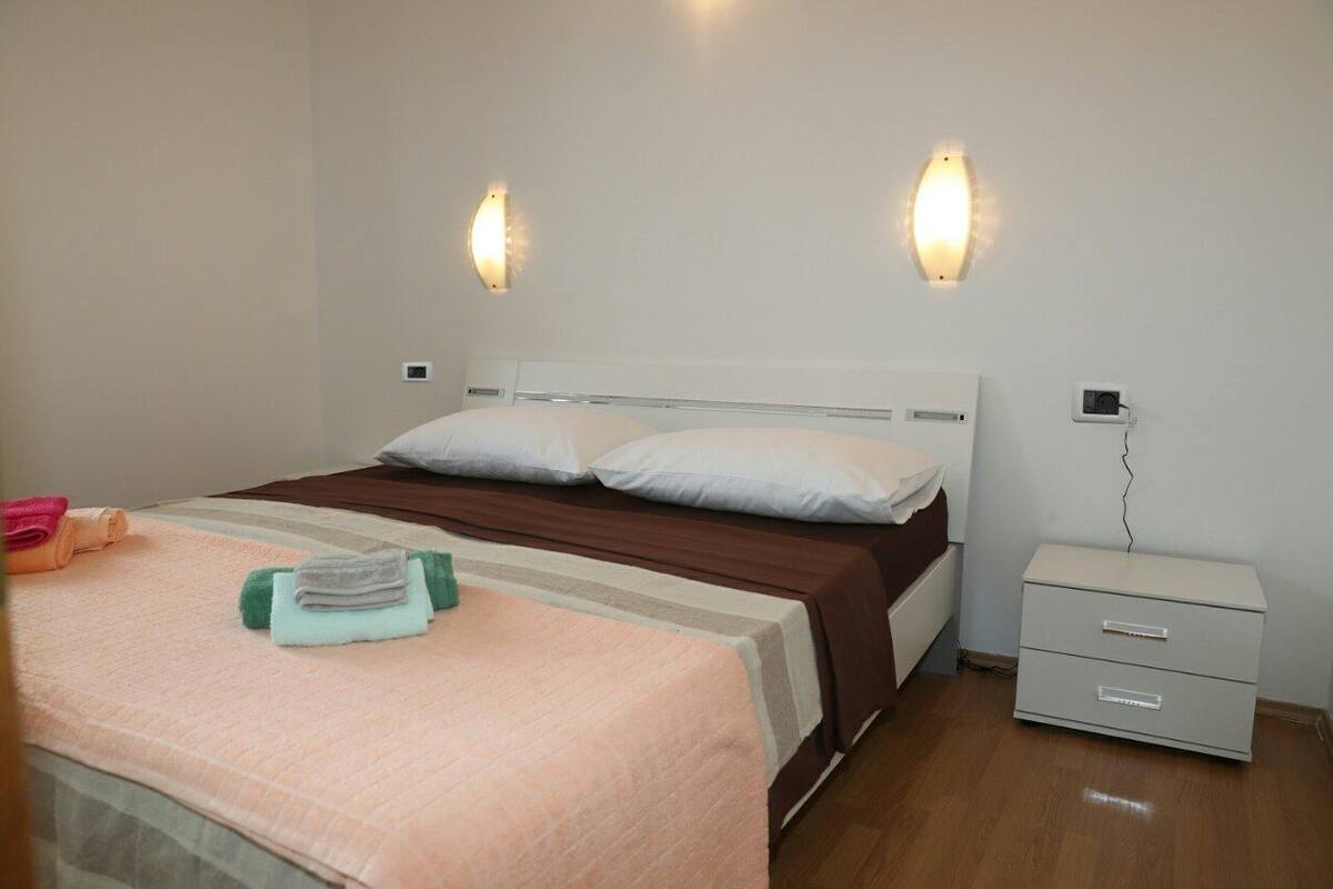 A neatly made bed with a brown headboard, two wall-mounted light fixtures, a bedside table with a power outlet, and a small white nightstand.