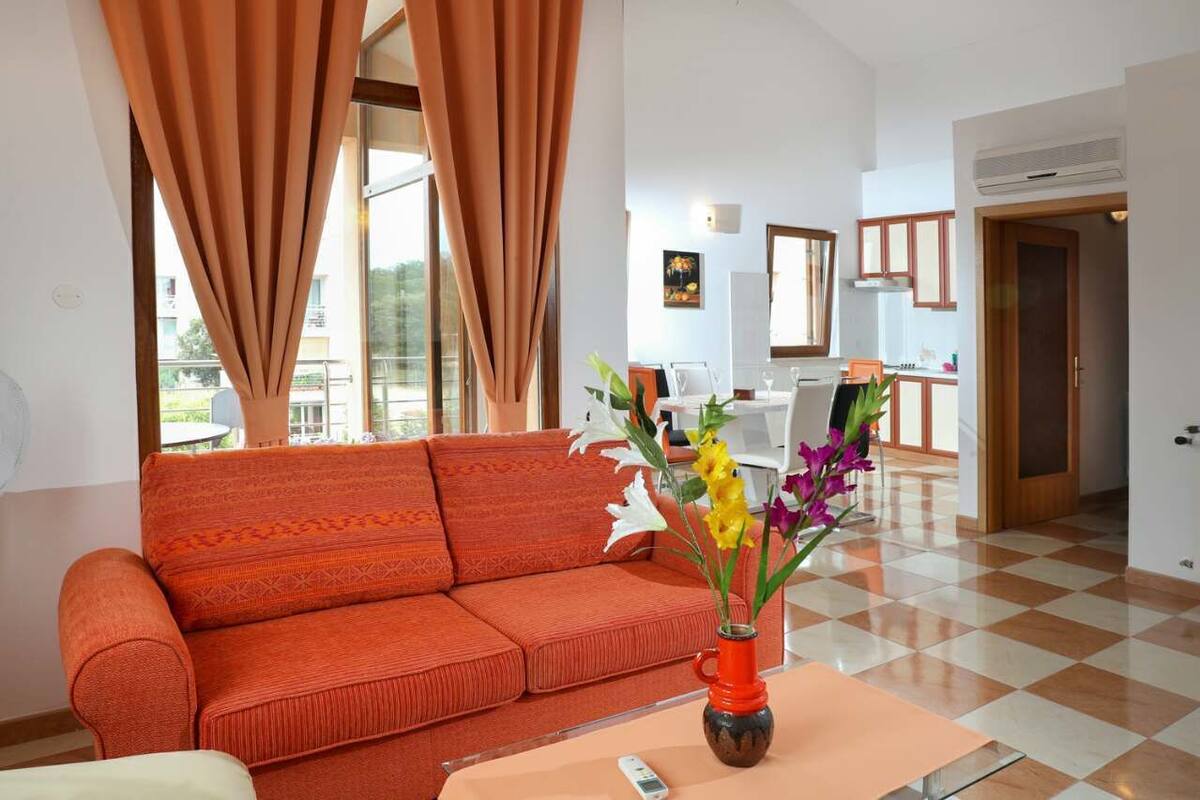 A modern living room with a sofa, a coffee table with a vase of flowers, and a view of the kitchen and balcony.