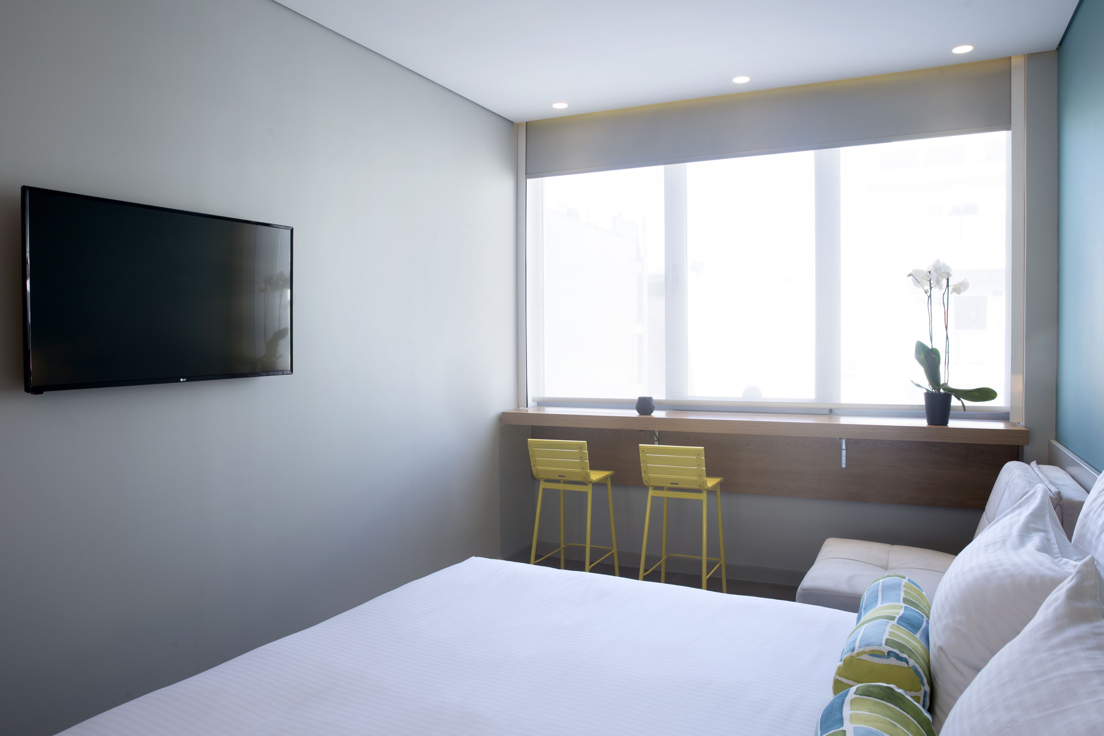 Et moderne hotelværelse med en stor seng, et fladskærms-tv monteret på væggen, to gule stole ved et vindue med byudsigt, og et lille bord med en potteplante.