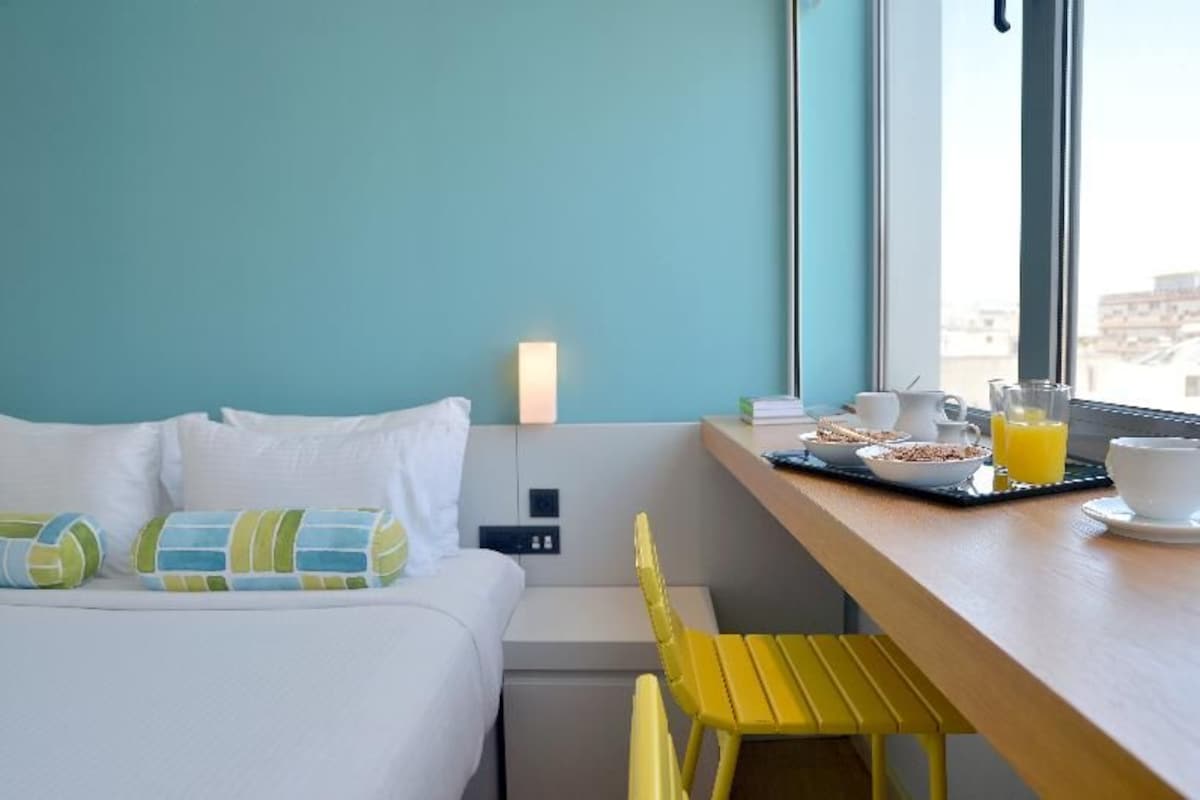 A hotel room with a bed, a small table with breakfast items, a chair, and a window with a city view.