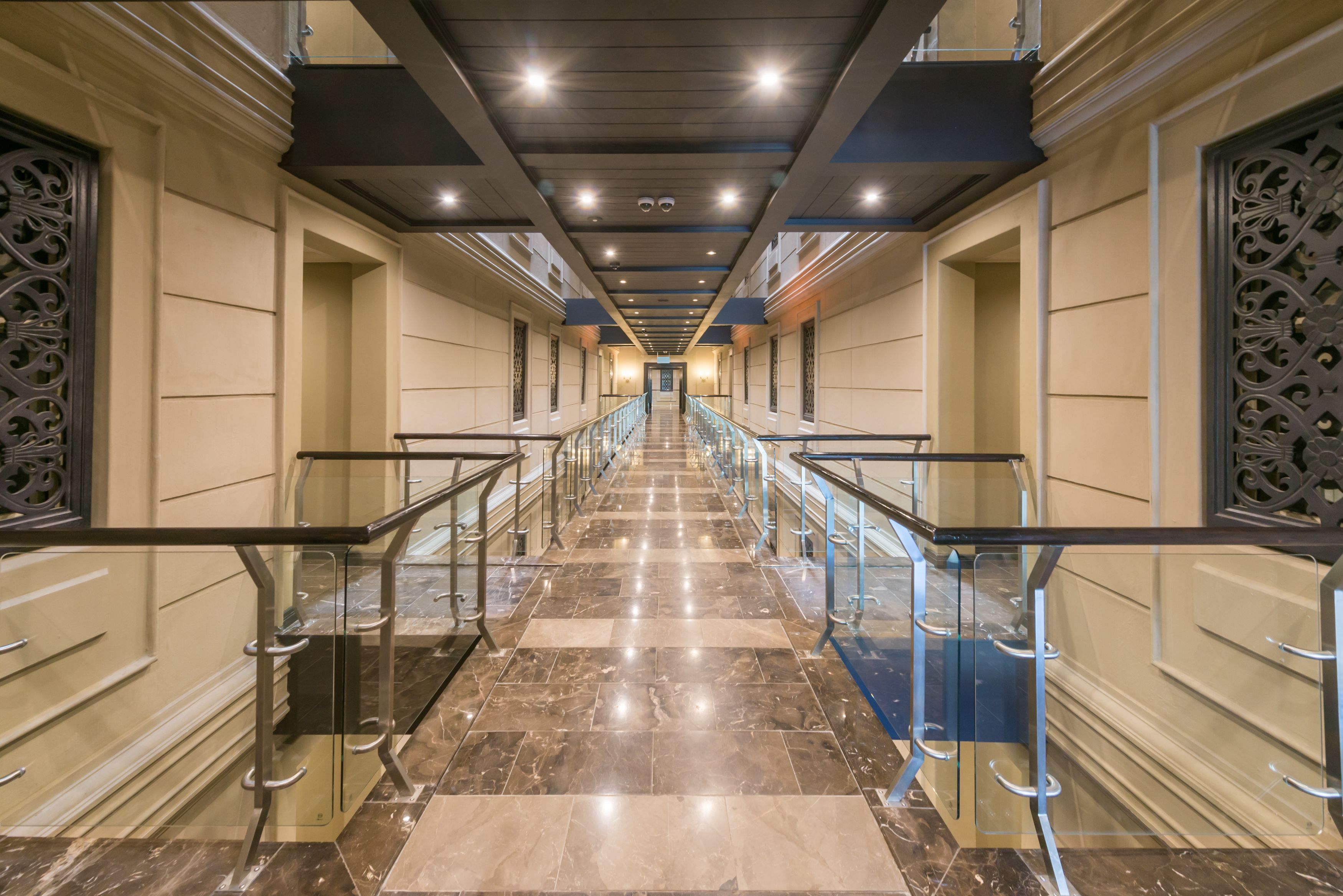 Un largo pasillo bien iluminado, con suelo de mármol y barandillas de vidrio, que conduce a una escalera.