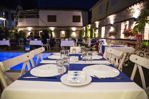 An outdoor dining area with tables set for two, white chairs, and a pool in the background. - Kervan Hotel (Antalya)