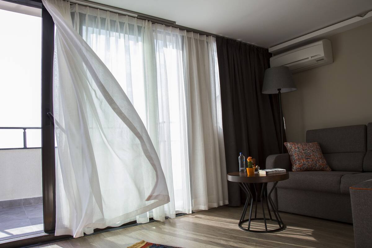 Una habitación con un sofá, una mesita con lámpara y una ventana con cortinas translúcidas.