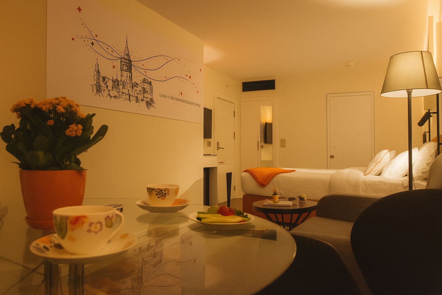 A cozy hotel room with a dining area, a bed with an orange throw, a standing lamp, and a wall with a cityscape illustration.