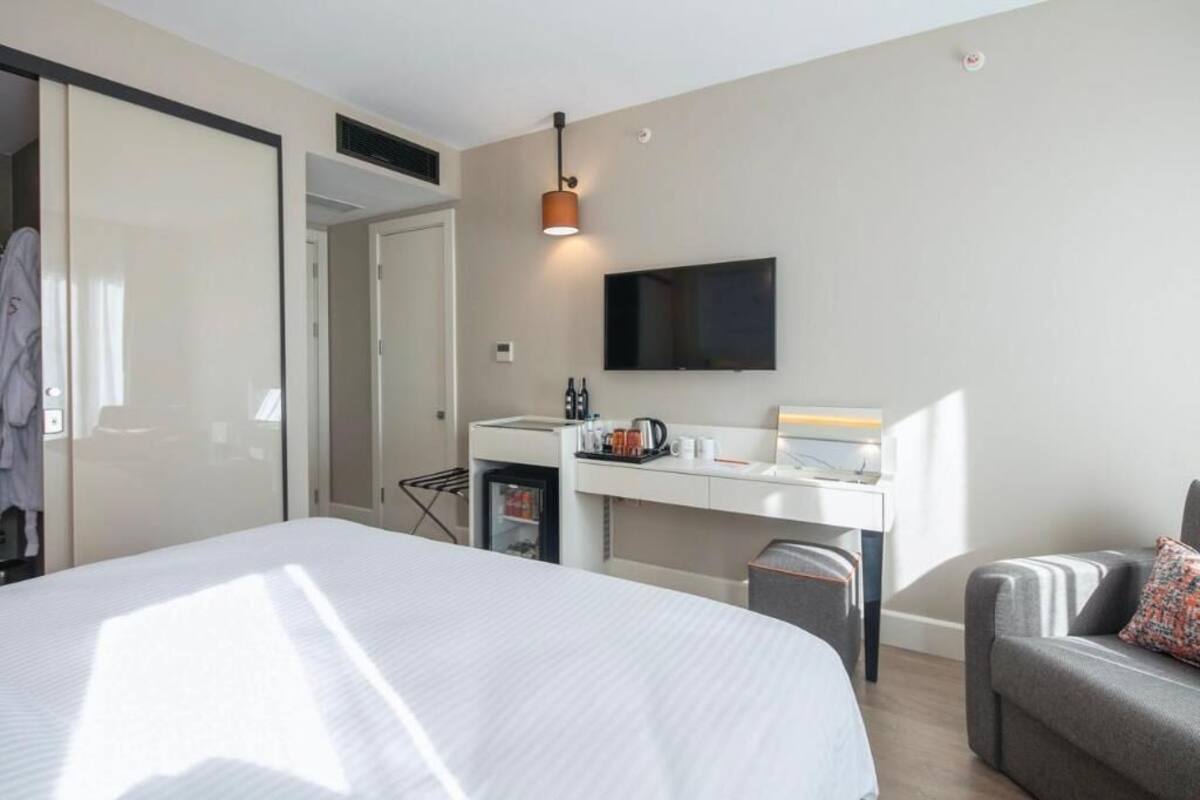 Habitación de hotel con cama, escritorio con televisor de pantalla plana, armario y zona de estar con cojín.