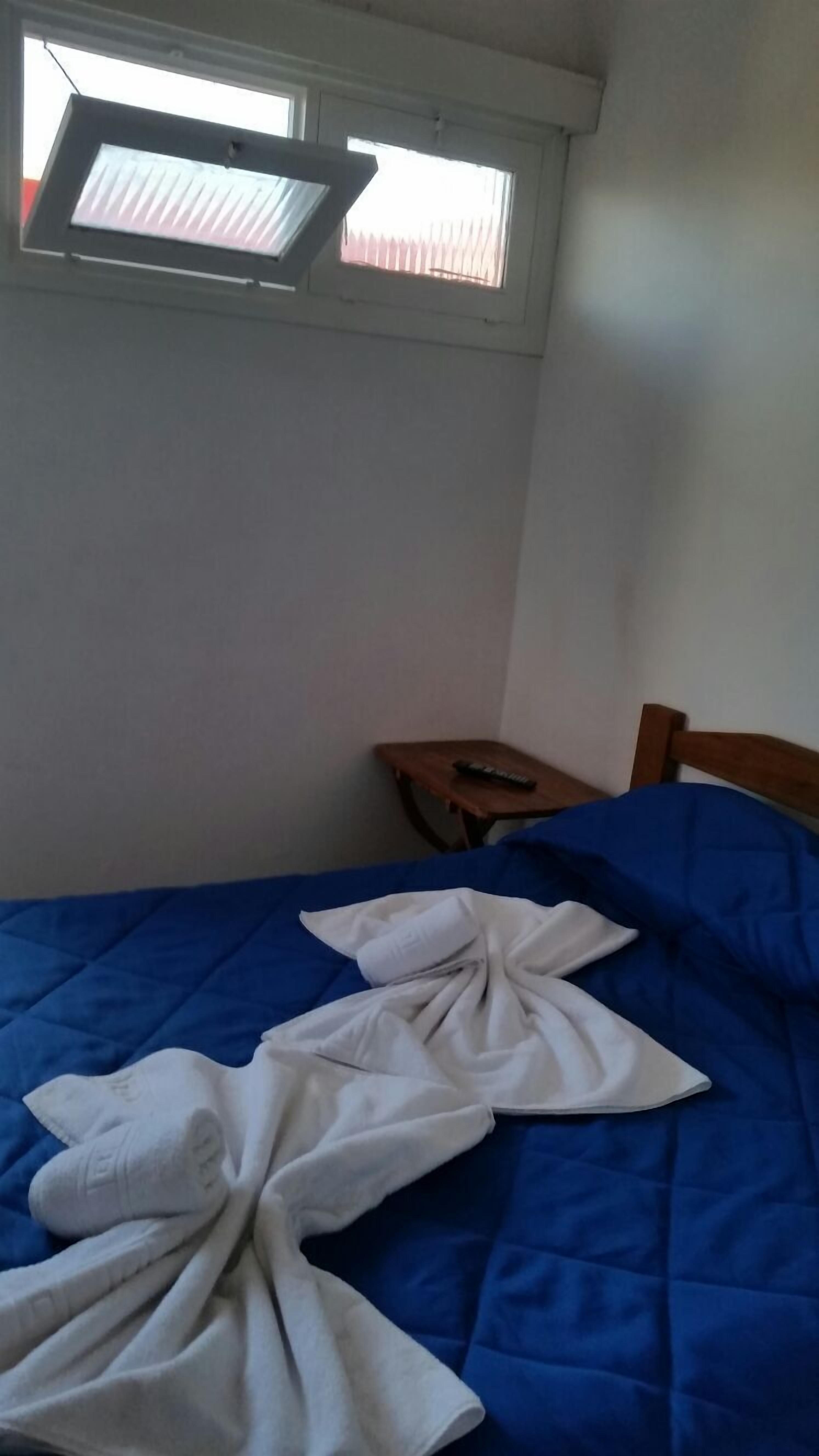 Una cama individual con cubierta azul, una mesita de noche de madera con un control remoto y una ventana sobre la cama.