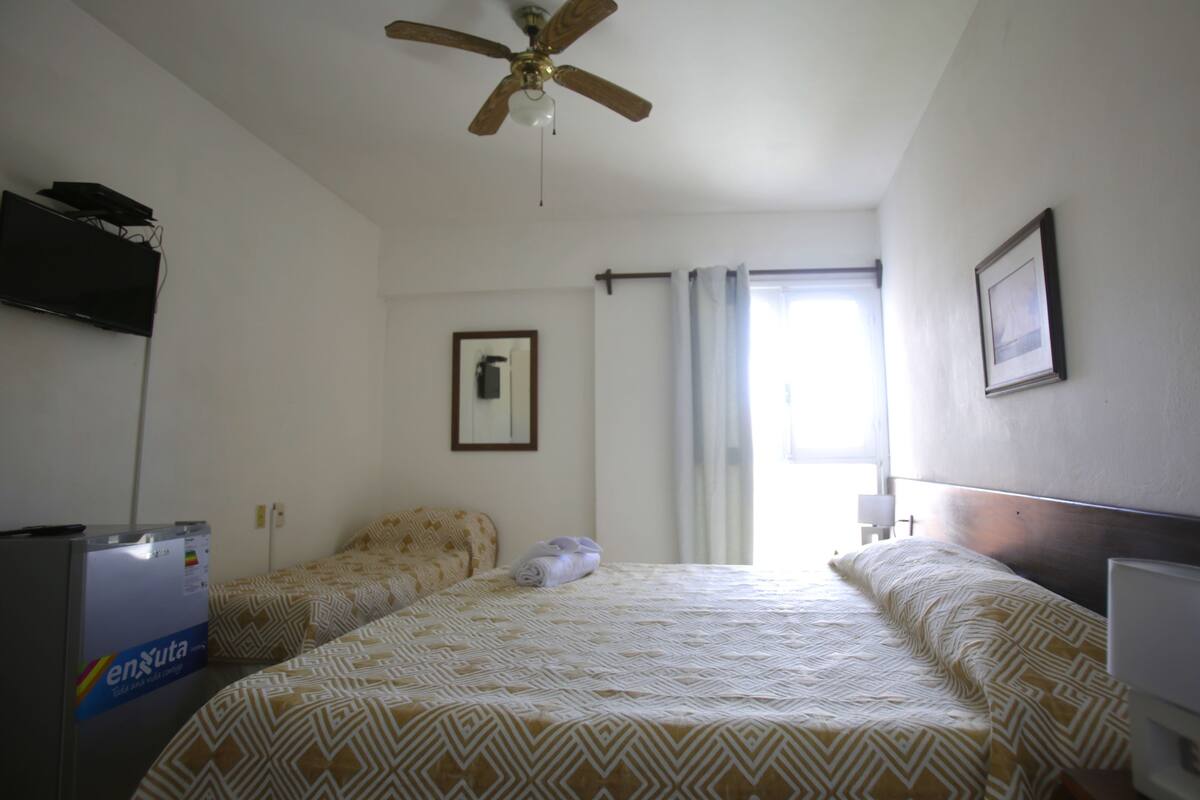 Ein Hotelzimmer mit zwei Betten, einem Deckenventilator, einem an der Wand montierten Fernseher und einem Fenster mit Vorhängen.