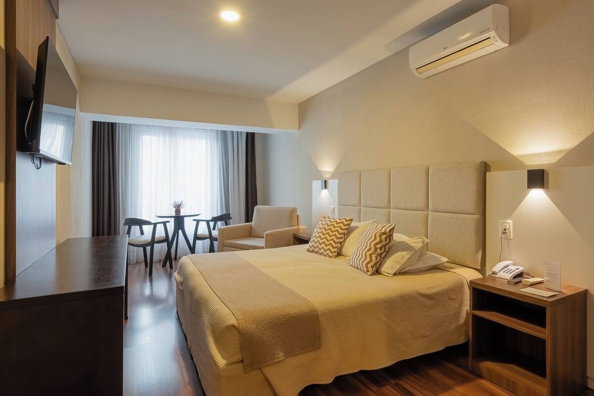 A hotel room with a bed, bedside table, lamp, TV, and a desk with chairs.