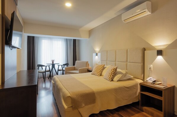 A hotel room with a bed, bedside table, lamp, TV, and a desk with chairs. - Hotel Querência (Gramado)