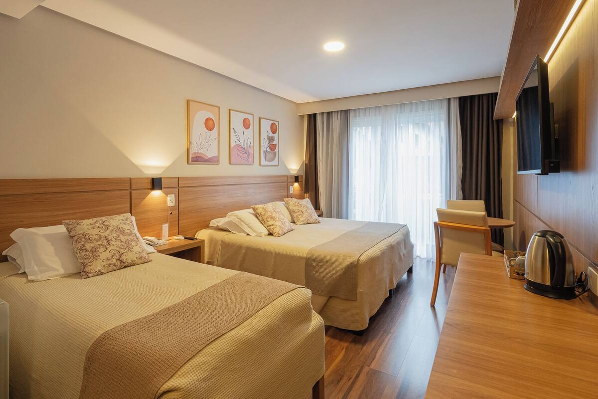 A hotel room with two beds, a wooden headboard, a desk with a kettle, and a TV mounted on the wall.