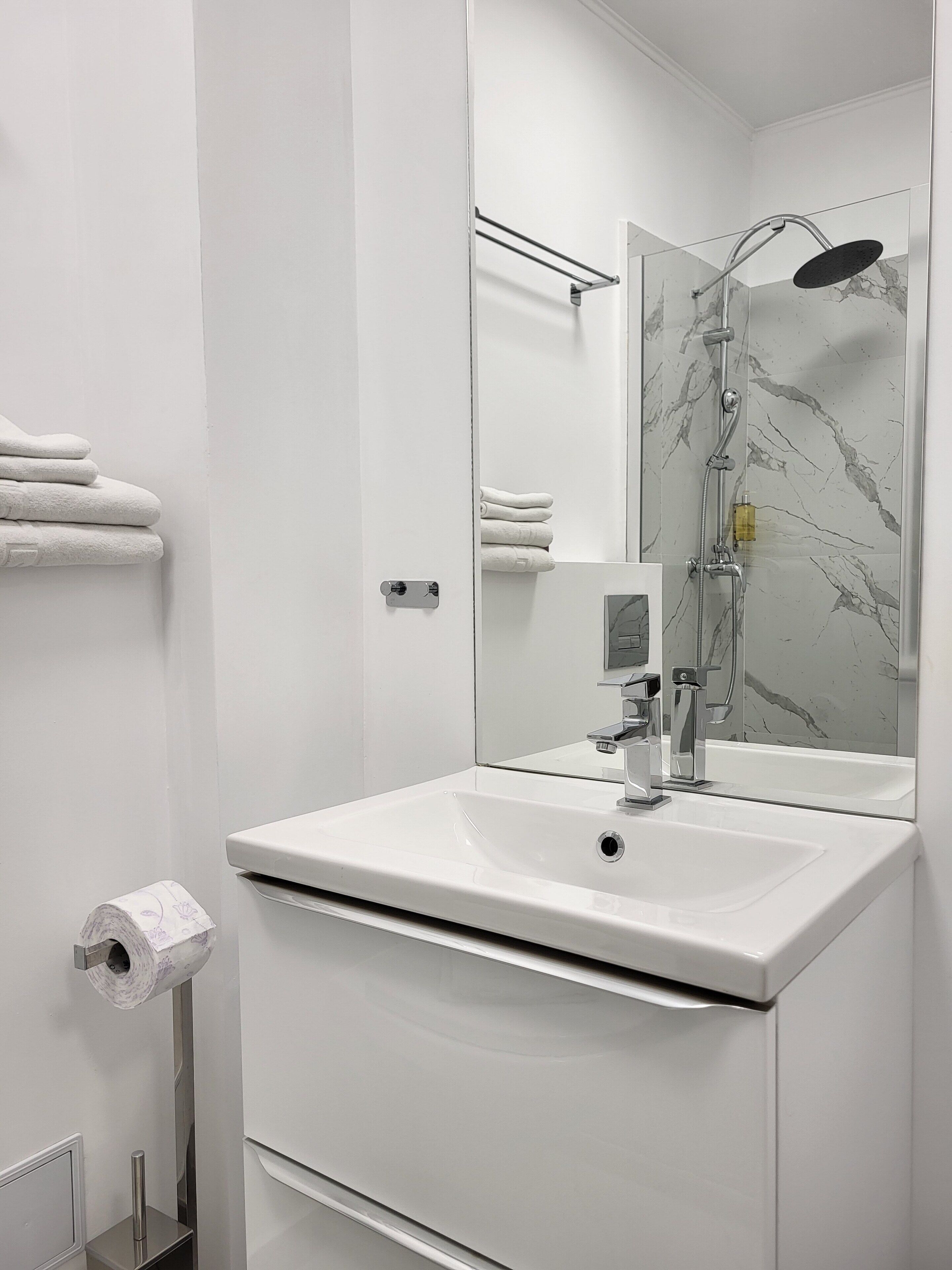 A modern bathroom with a white sink, a mirror, a shower, and neatly folded towels on a rack.