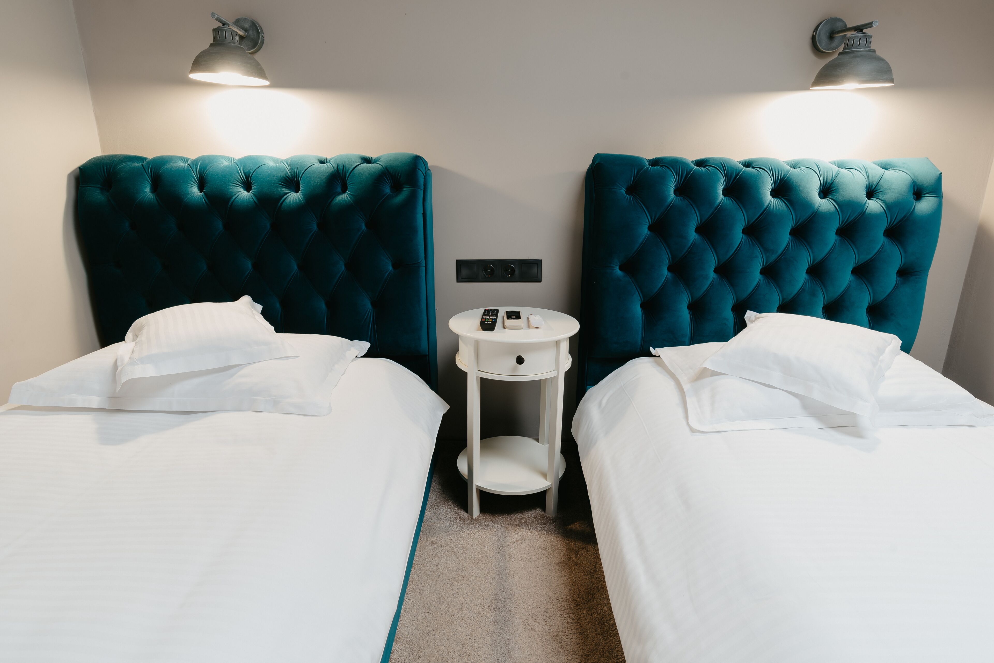 A hotel room with two beds, each with a teal headboard, white bedding, and a small bedside table with a lamp.