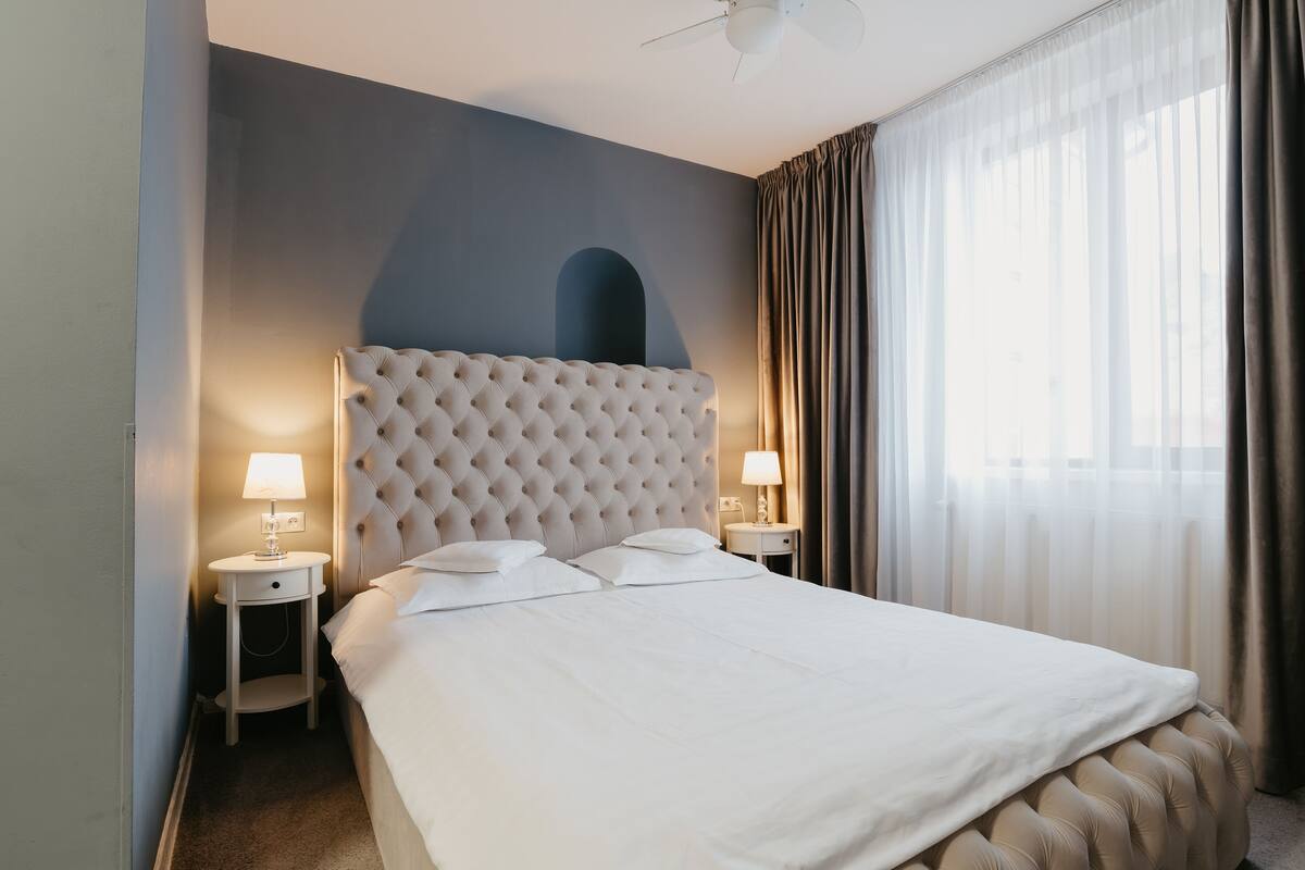 A neatly made bed with a tufted headboard, two bedside tables with lamps, and a large window with sheer curtains.