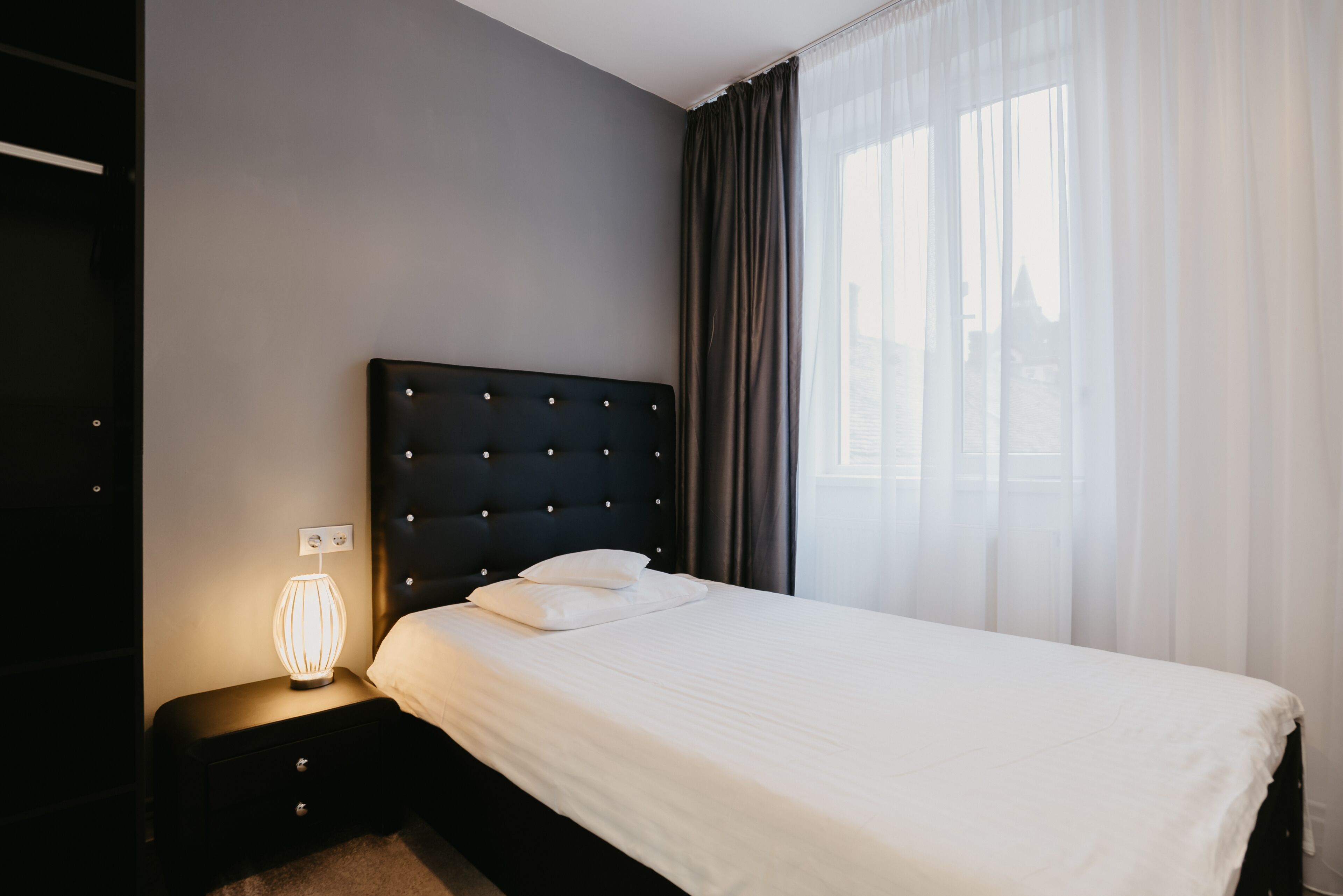 Un lit bien fait, avec une tête de lit capitonnée, une lampe de chevet et une fenêtre ornée de rideaux légers.