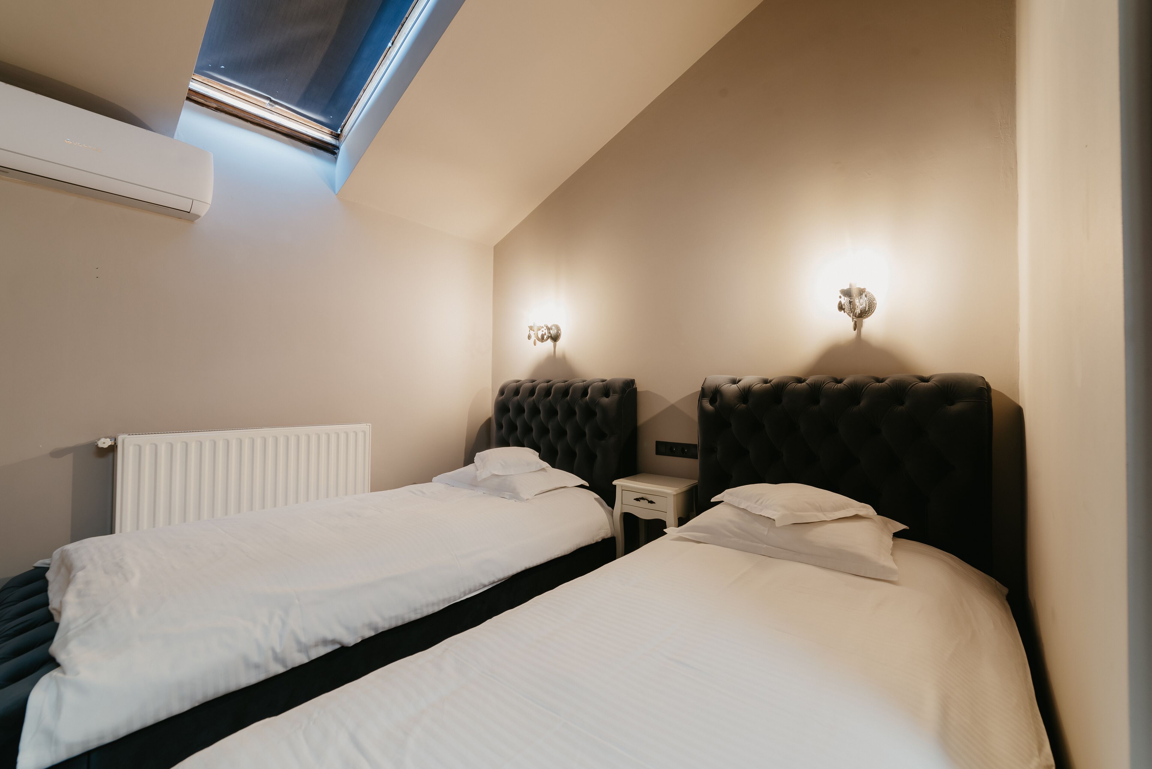 A bedroom with two beds, a radiator, a small bedside table, and a wall-mounted light fixture.