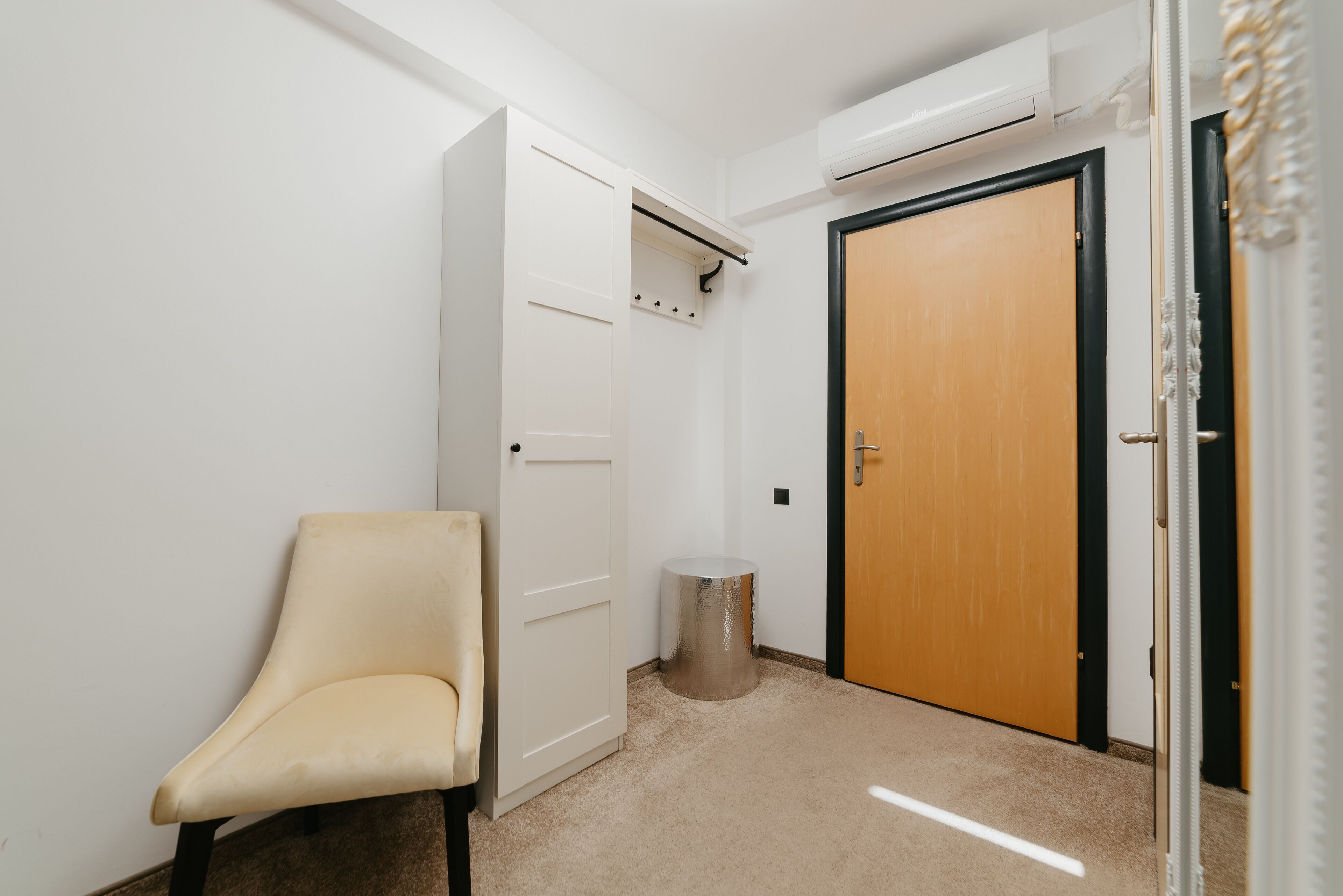 A hallway with a beige chair, a white wardrobe, a wooden door, and a trash bin.