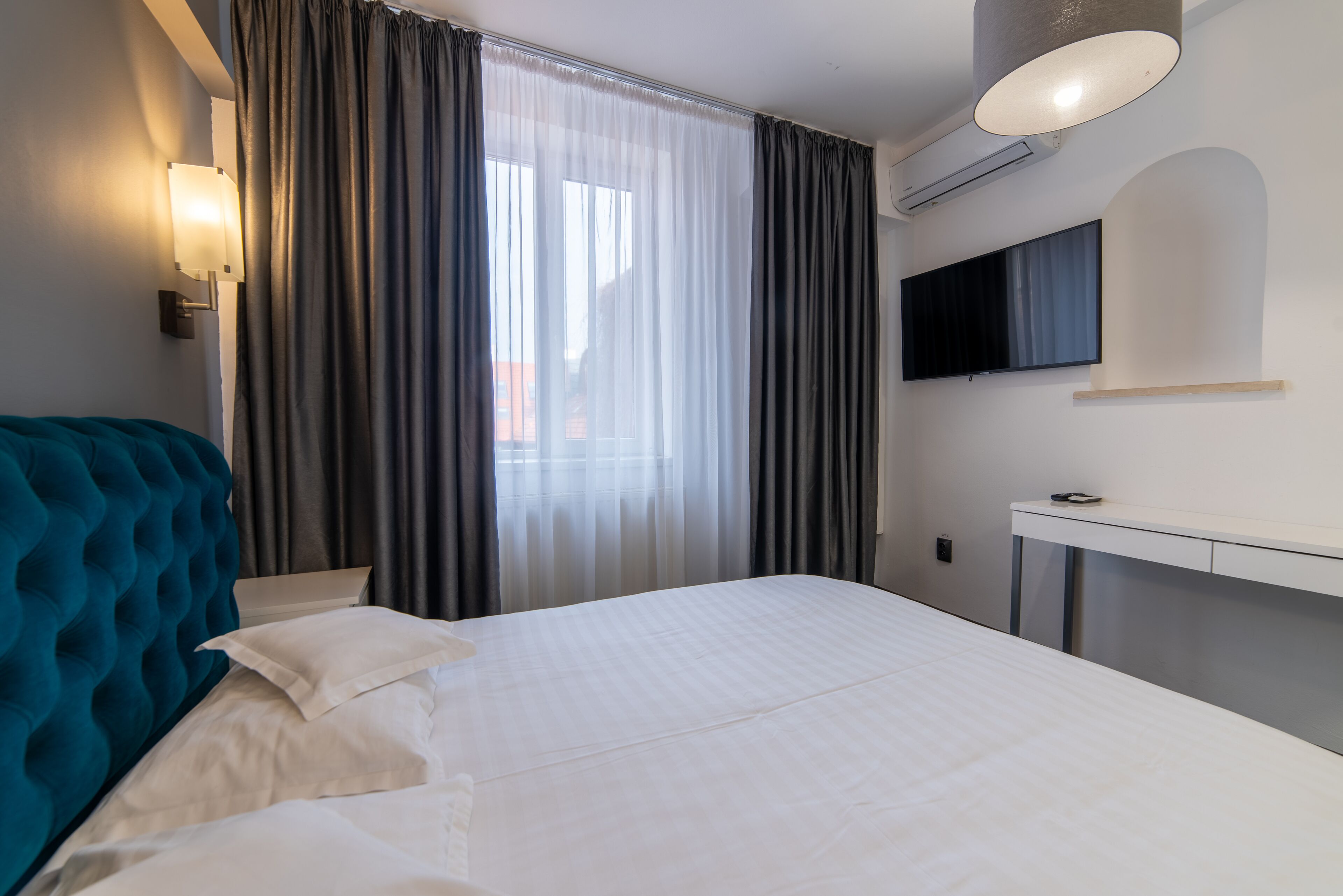 Habitación de hotel con una cama grande, cabecera azul, mesita de noche, televisor de pantalla plana y ventana con cortinas.