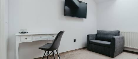 Una habitación con un escritorio blanco, una silla negra, un sofá gris y un televisor de pantalla plana montado en la pared.