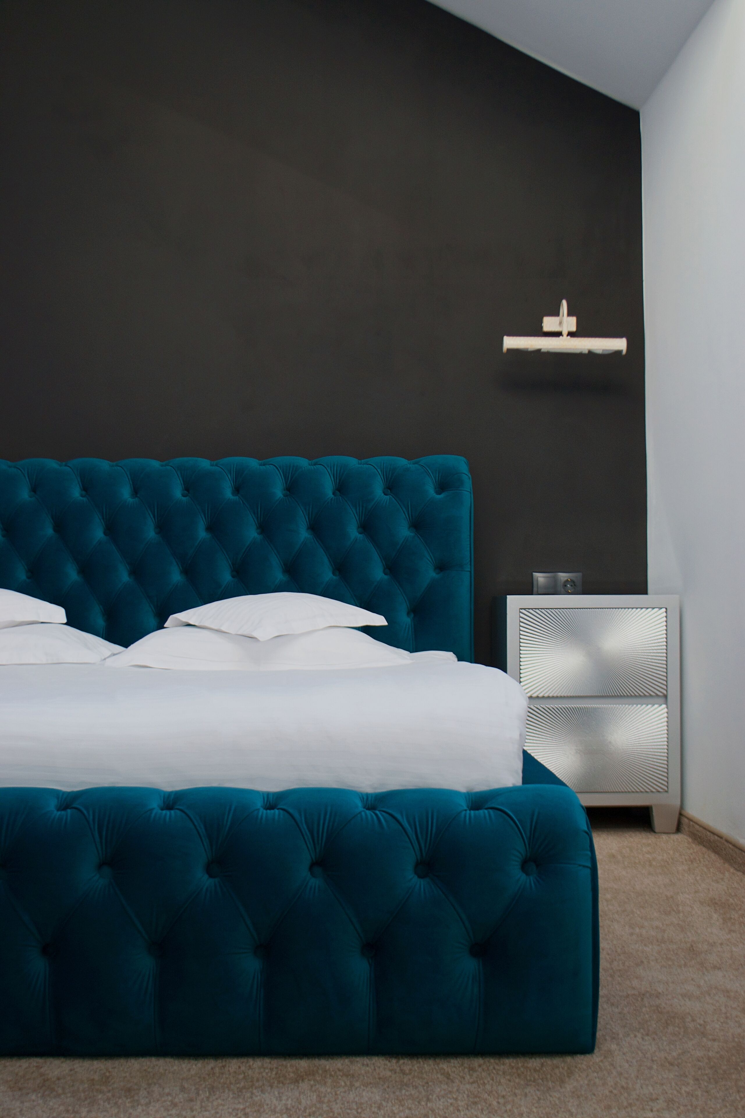 Une chambre à coucher avec une tête de lit turquoise à motifs capitonnés, une literie blanche, une table de chevet et une étagère fixée au mur.