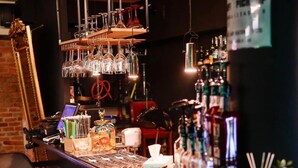 A bar area with a wooden counter, hanging glasses, and a variety of bottles. There are framed pictures on the wall and a sign that says "PICK UP." - Fullton Central Inn (Cluj-Napoca)