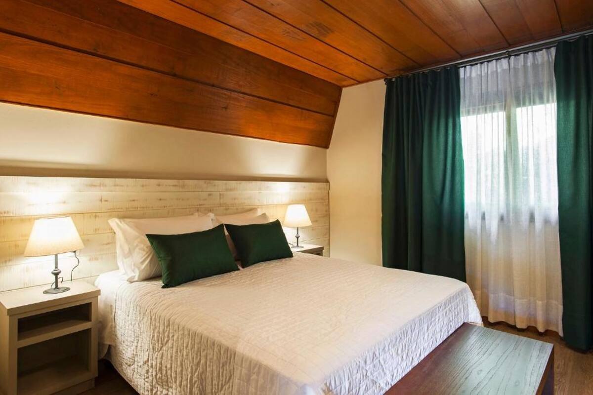 A room with a wooden ceiling, a bed with white bedding, two bedside lamps, and a window with green curtains.