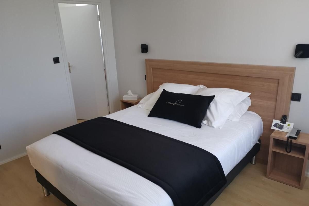 Un lit bien fait, avec une couvre-lit noir et blanc, une tête de lit en bois, une table de chevet avec un téléphone et une lampe fixée au mur.