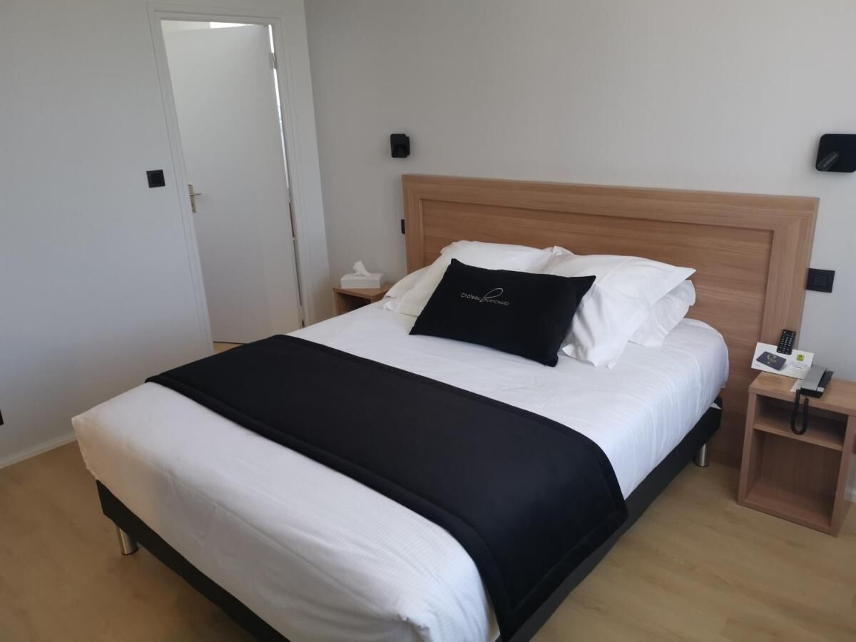 Una cama bien hecha con colcha blanco y negro, un cabecero de madera, una mesita de noche con un teléfono y una lámpara fijada a la pared.