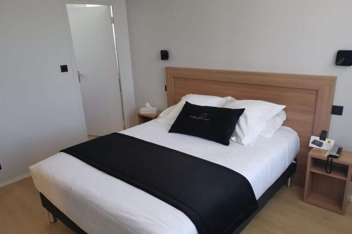 Un lit bien fait, avec une couvre-lit noir et blanc, une tête de lit en bois, une table de chevet avec un téléphone et une lampe fixée au mur.
