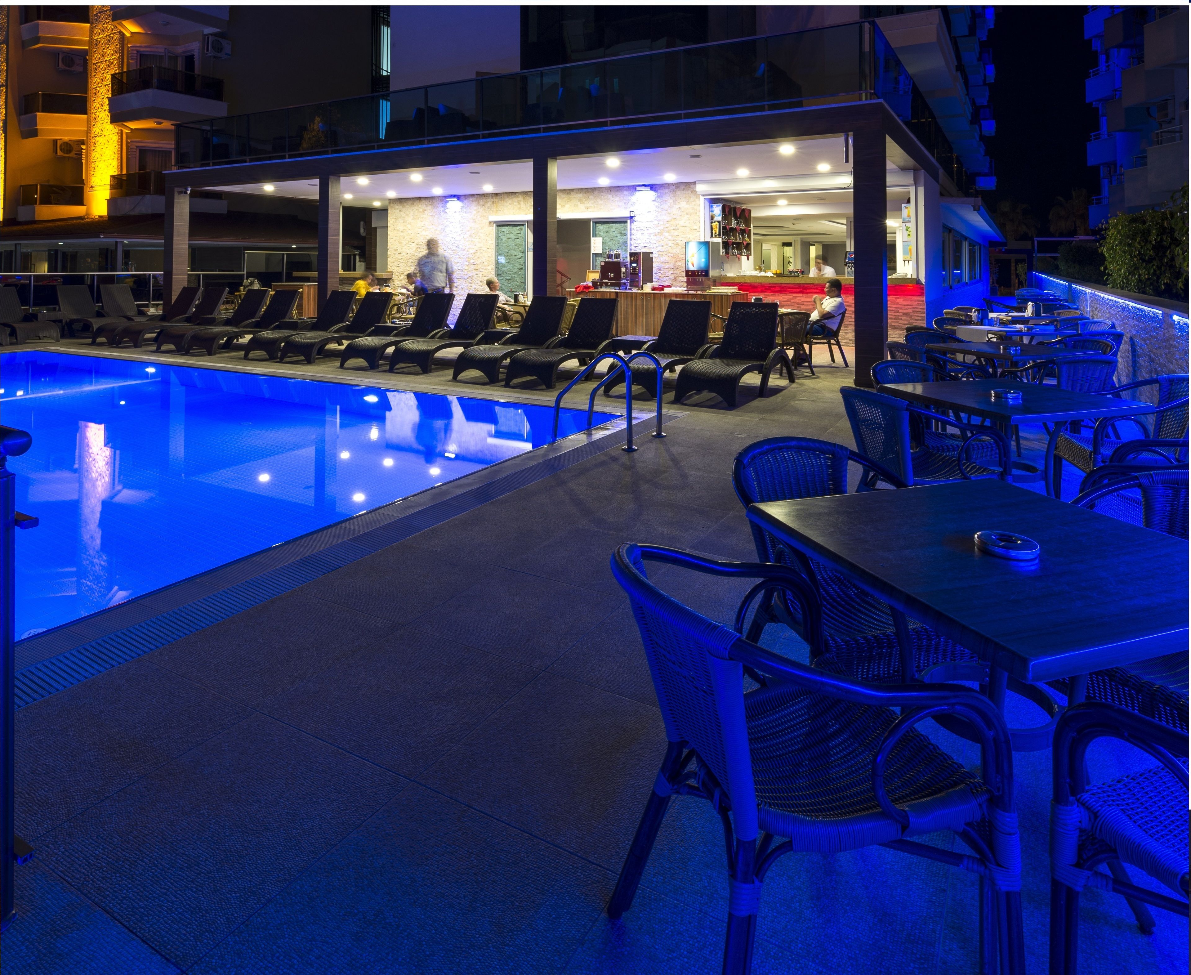 Un área junto a la piscina con sillones de descanso y mesas, una zona de bar y una piscina con iluminación azul.