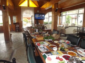 A restaurant with a long dining table set with various dishes, a television screen displaying a menu, and a view of the outdoors through large windows.