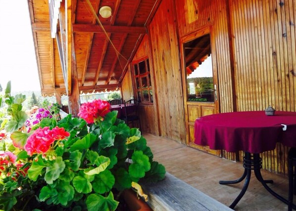 A wooden building with a covered porch, a table with a red tablecloth, and potted plants with pink flowers.