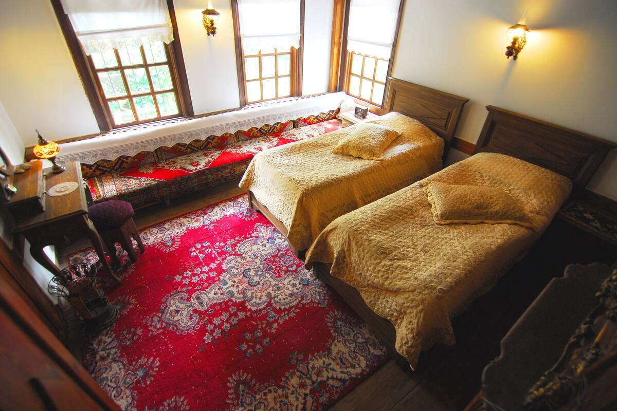 A traditional room with two beds, a red patterned carpet, and a window with a view of greenery.