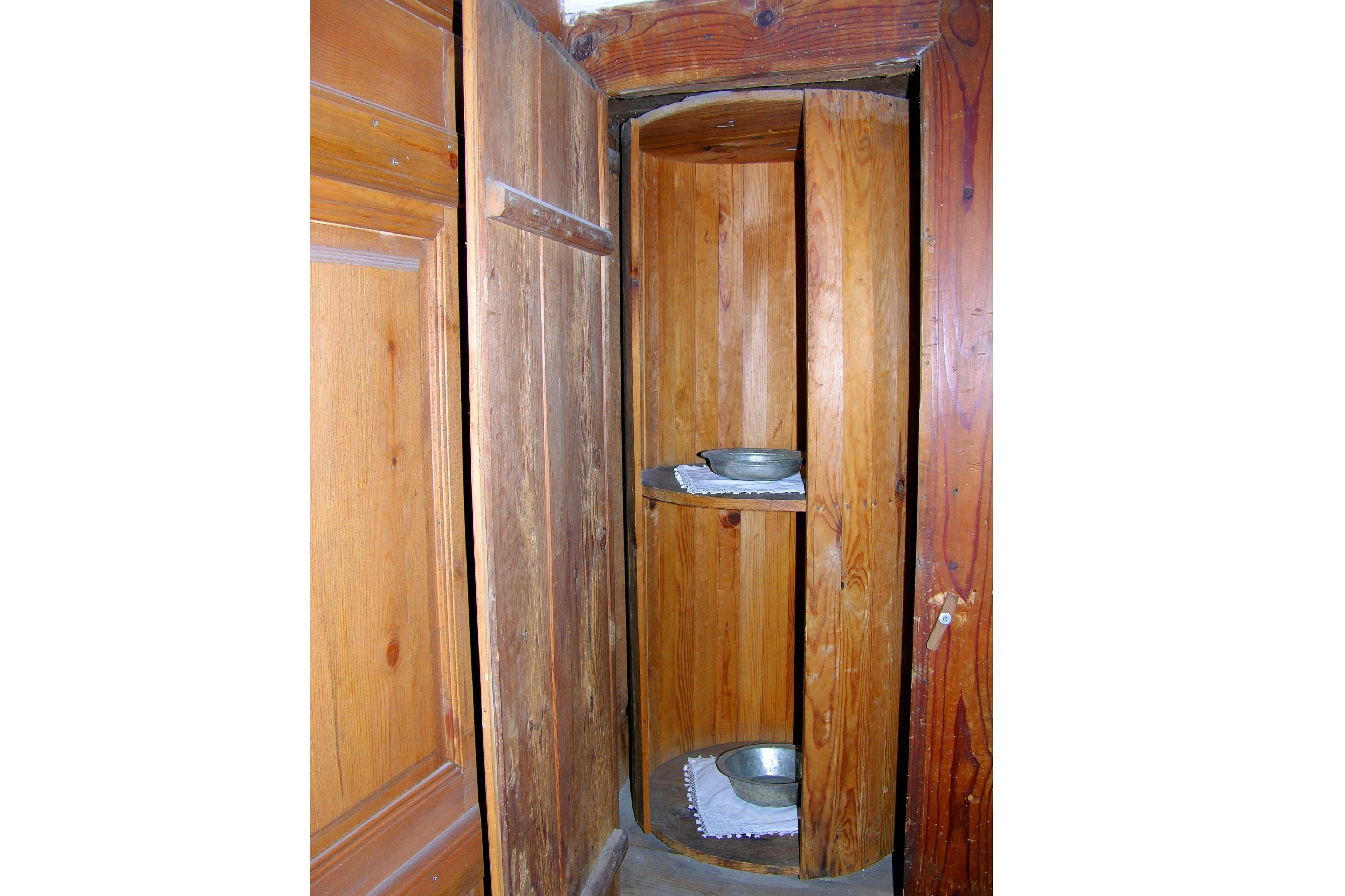 Cabina de baño de madera con un inodoro metálico en el suelo y un recipiente metálico en un estante.