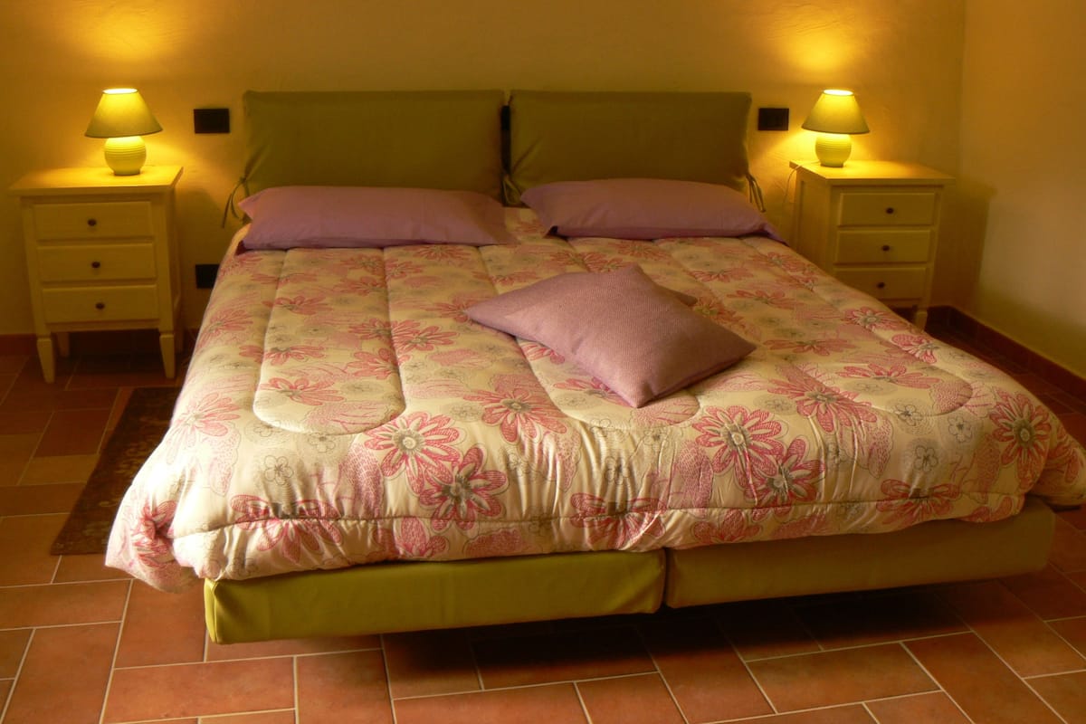 Uma cama com colcha florida, duas mesas de cabeceira com luminárias e um painel de cabeceira verde.