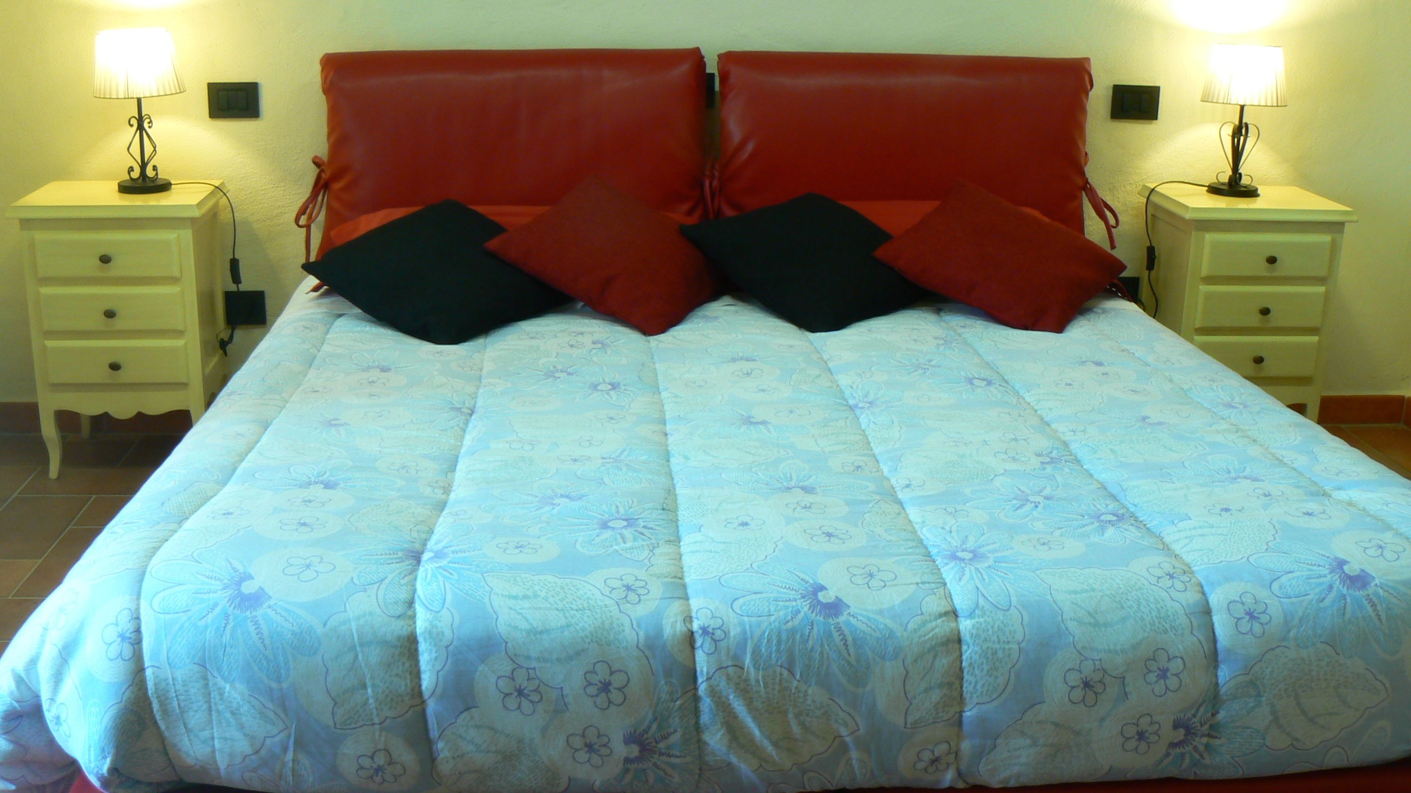 Un lit avec une couette à motifs floraux, deux oreillers noirs et une tête de lit rouge. Il y a deux tables de chevet, chacune avec une lampe, de chaque côté du lit.