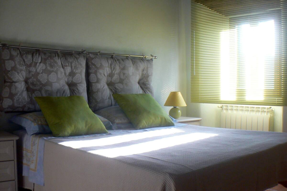 Cama bem arrumada com cabeceira, dois travesseiros verdes, abajur e janela com persianas.