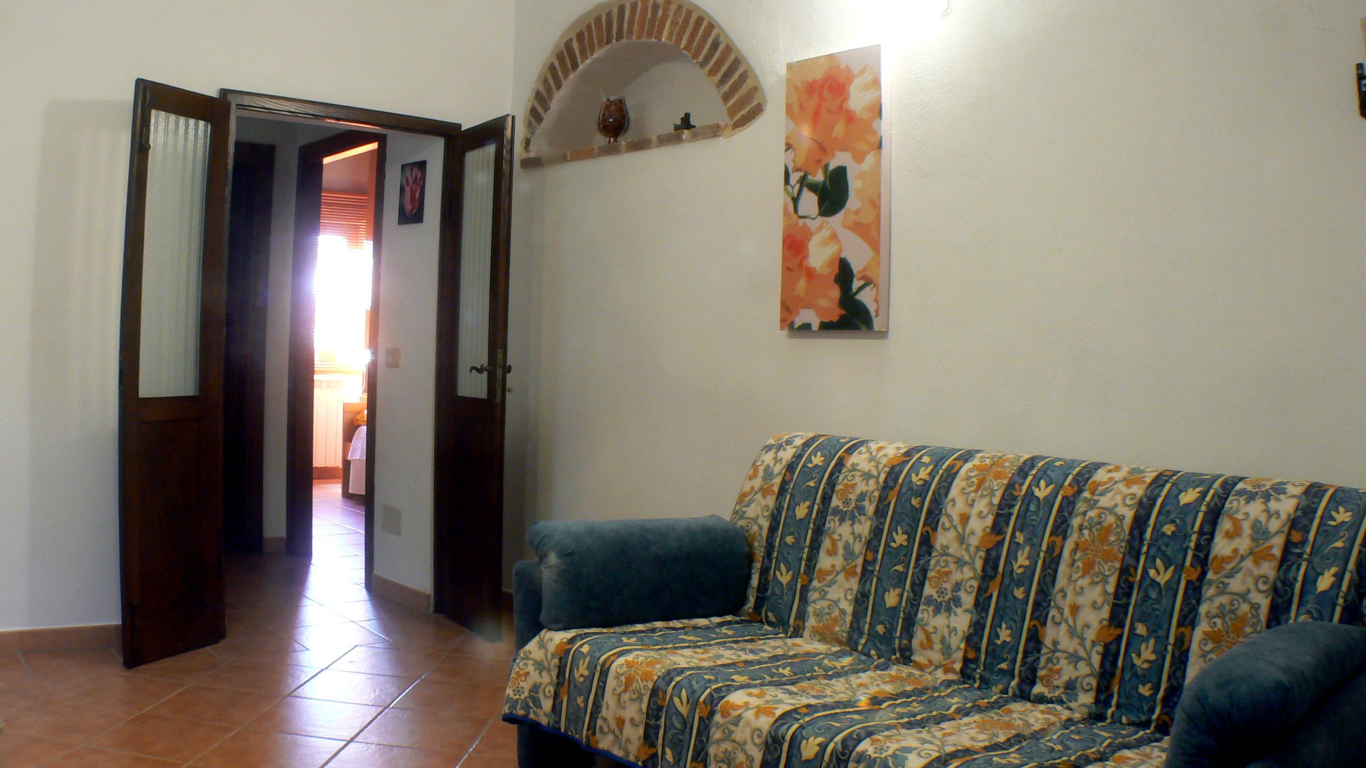 A living room with a patterned sofa, a tiled floor, a doorway leading to another room, and a wall-mounted shelf with decorative items.