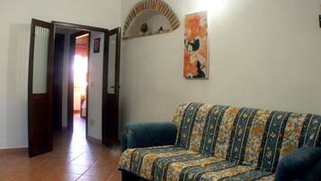 A living room with a patterned sofa, a tiled floor, a doorway leading to another room, and a wall-mounted shelf with decorative items.