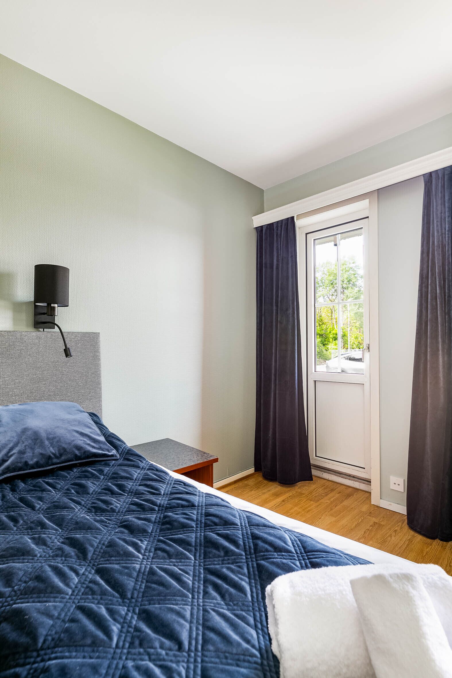 A bedroom with a bed, a nightstand, a wall-mounted lamp, a sliding glass door, and a view of greenery outside.