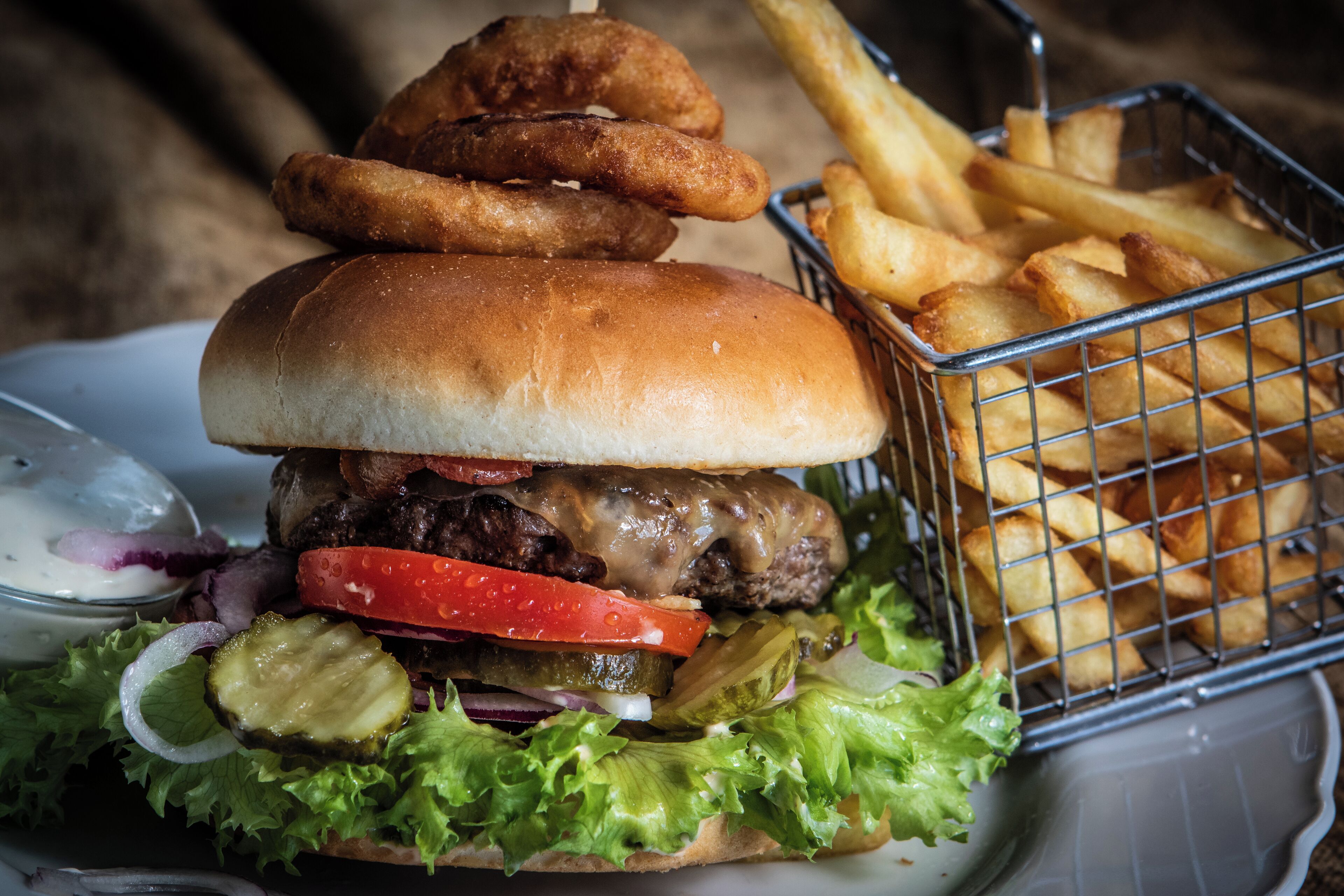 A burger with a bun, lettuce, tomato, and pickles, accompanied by a basket of fries and onion rings.