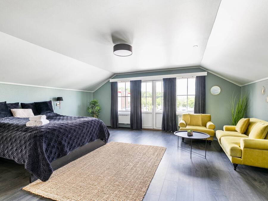Un dormitorio moderno con cama, un sillón amarillo, un sofá, una mesita y un amplio ventanal con cortinas.