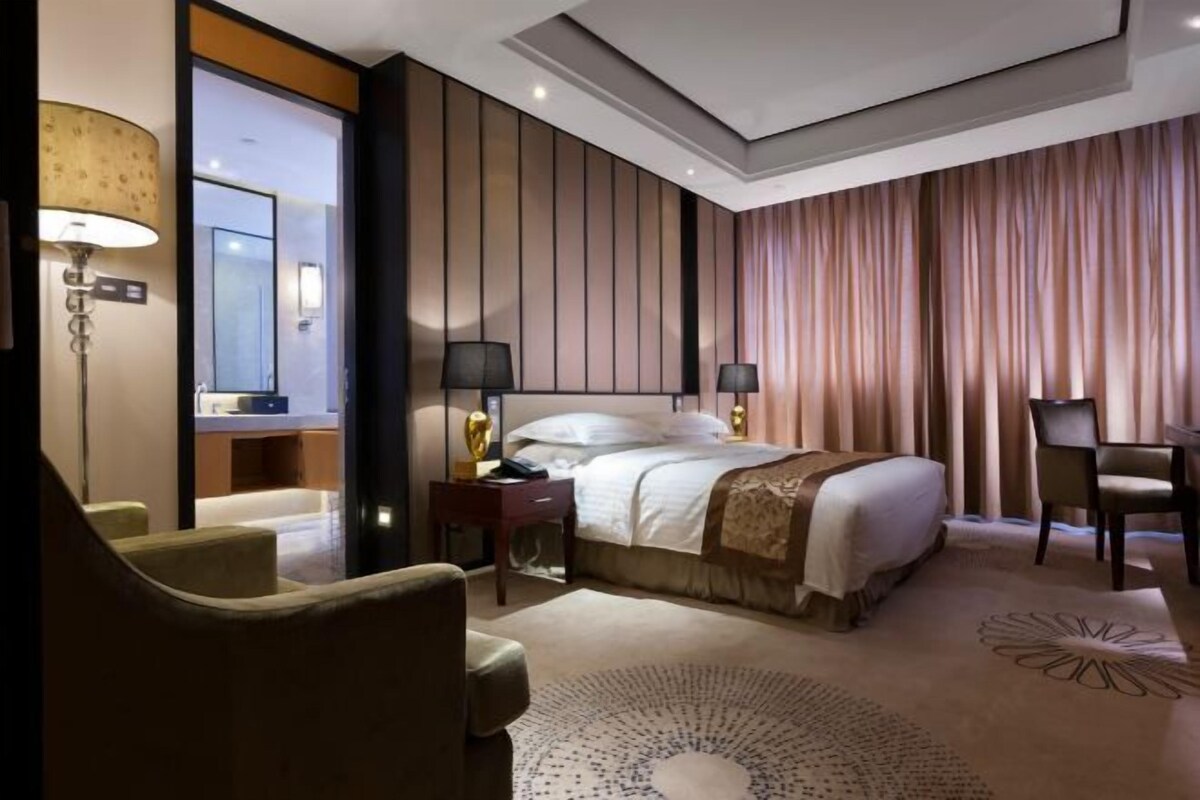 A hotel room with a bed, two bedside lamps, a desk, a chair, a lamp, and a mirror.