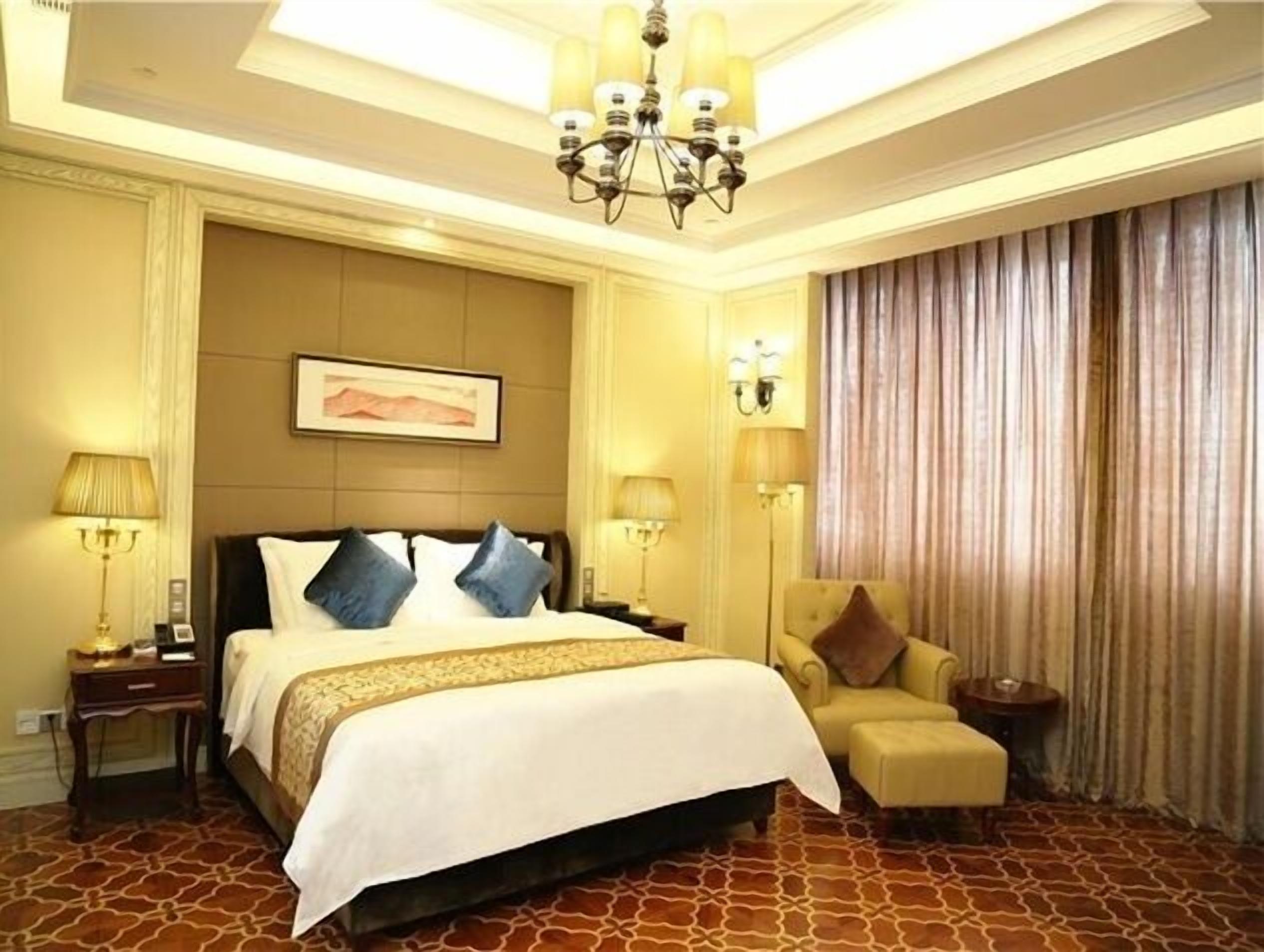 A hotel room with a large bed, a chair, a chandelier, and a painting on the wall.