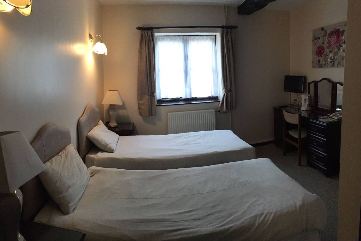 A hotel room with two single beds, a desk with a chair, a lamp, a radiator, and a window with curtains.