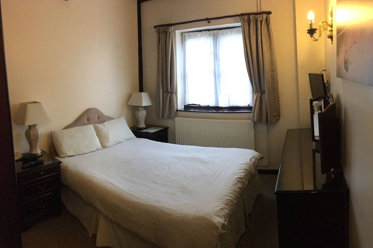 A neatly made bed in a hotel room with a window, a bedside table with a lamp, and a dresser.