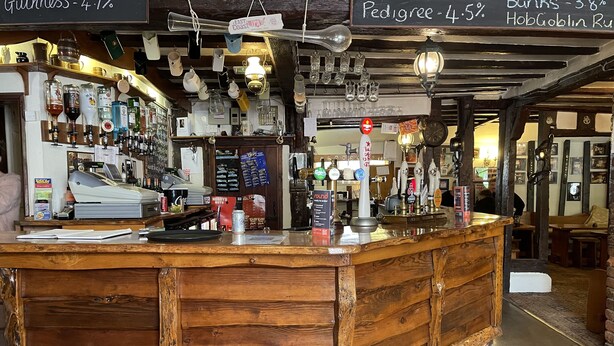 Una zona de bar rústica con mostrador de madera, varias botellas y un menú en pizarra que muestra precios.