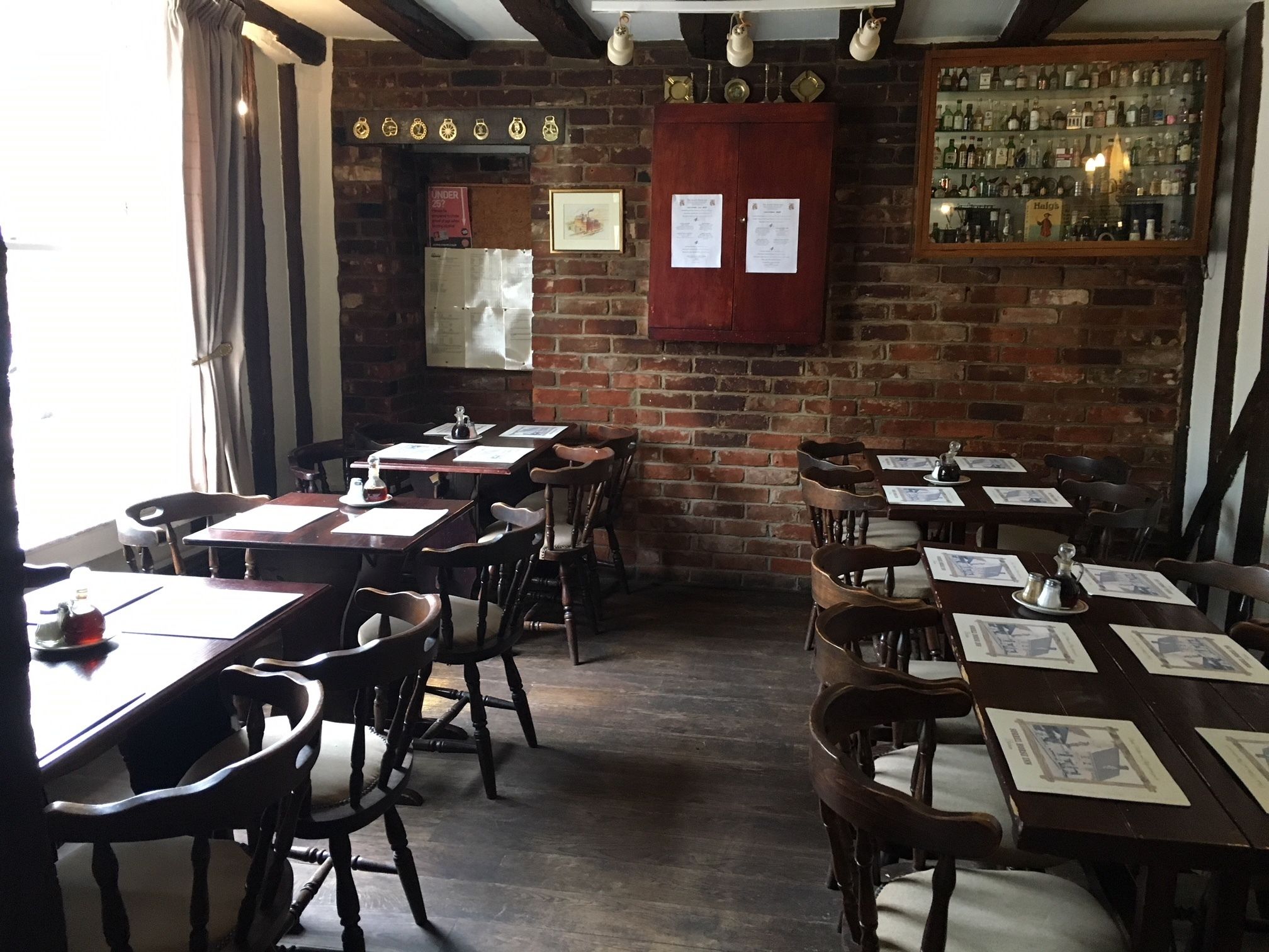 A restaurant with brick walls, wooden tables, and chairs. There are menus on the tables, and a bar area with bottles visible.
