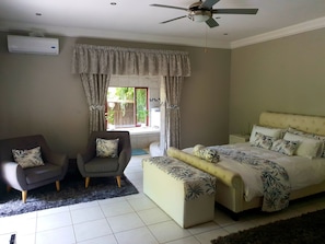 A bedroom with a bed, two armchairs, a ceiling fan, and a window with curtains.