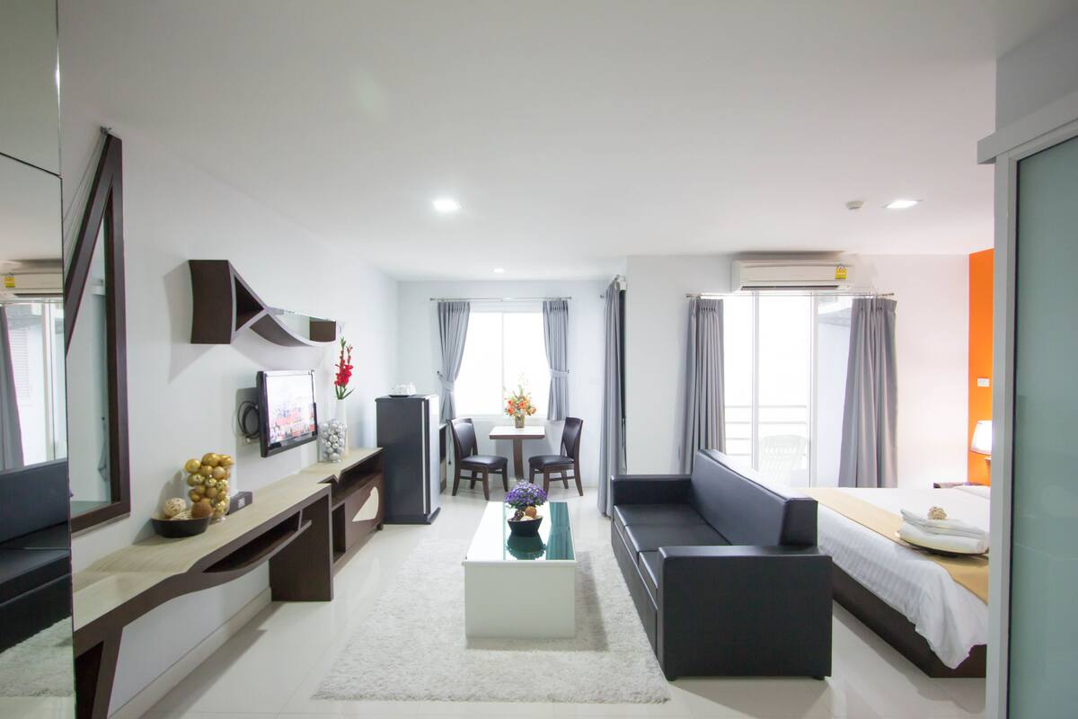 A modern hotel room with a sofa, a dining table, a TV, and a large window.