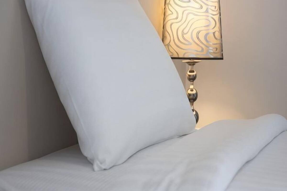 A bed with a white pillow and a wall-mounted lamp with a patterned shade.